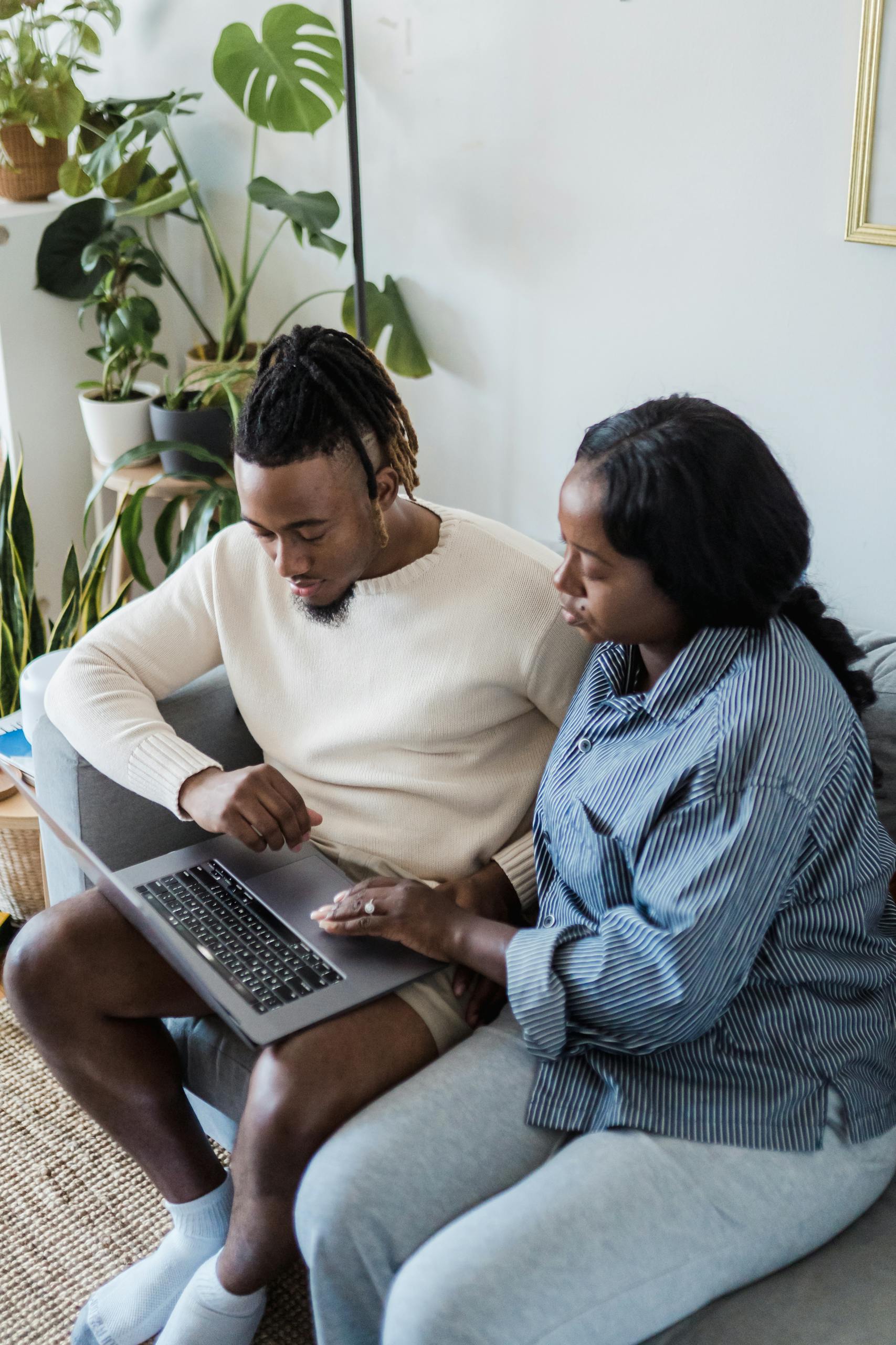 A couple sits on a sofa using a laptop in a cozy living room surrounded by indoor plants.